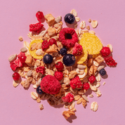 Eine Müsli-Mischung auf rosa Hintergrund mit gefriergetrockneten Himbeeren, Blaubeeren, gelben Fruchtstücken und Haferflocken.