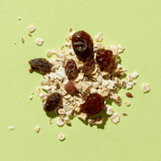 Eine Nahaufnahme der Bircher Müsli-Mischung auf hellgrünem Grund mit Haferflocken, Rosinen und getrockneten Apfelstücken.