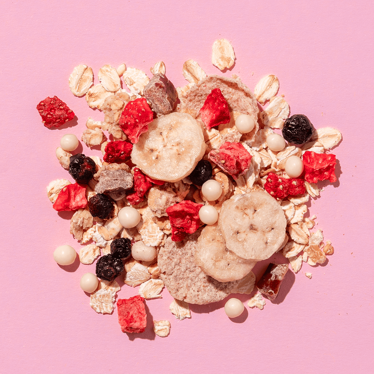 Eine lose Mischung aus Müsli-Zutaten auf rosa Grund: Haferflocken, getrocknete Erdbeerstücke, Bananenscheiben und dunkle Beeren.
