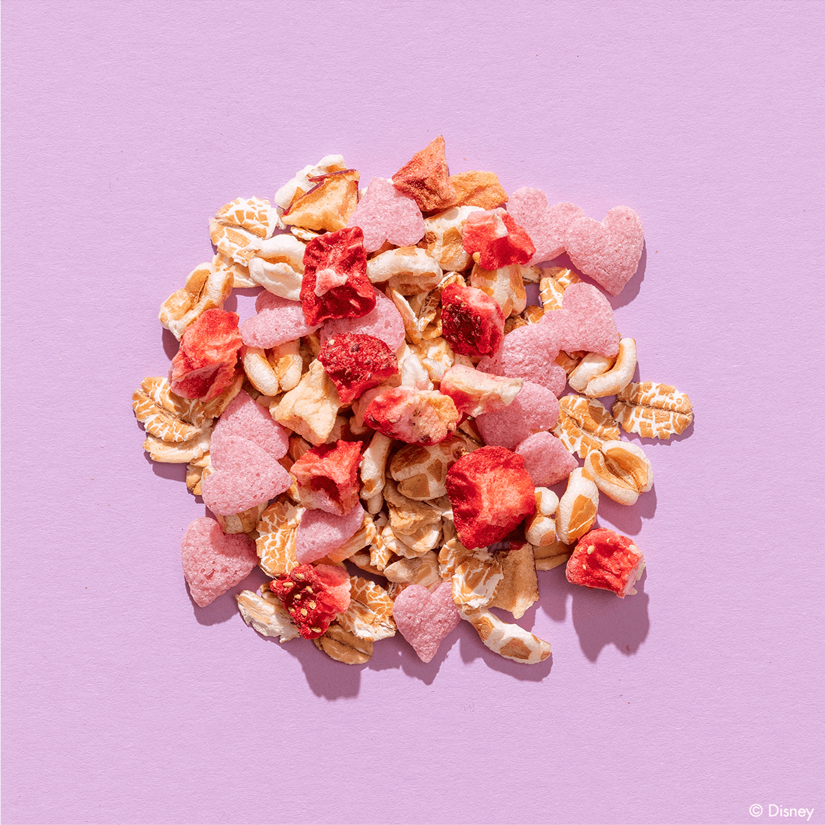 Eine bunte Müsli-Mischung auf violettem Hintergrund, die kleine rosa Schoko-Herzen und getrocknete Erdbeerstücke enthält.