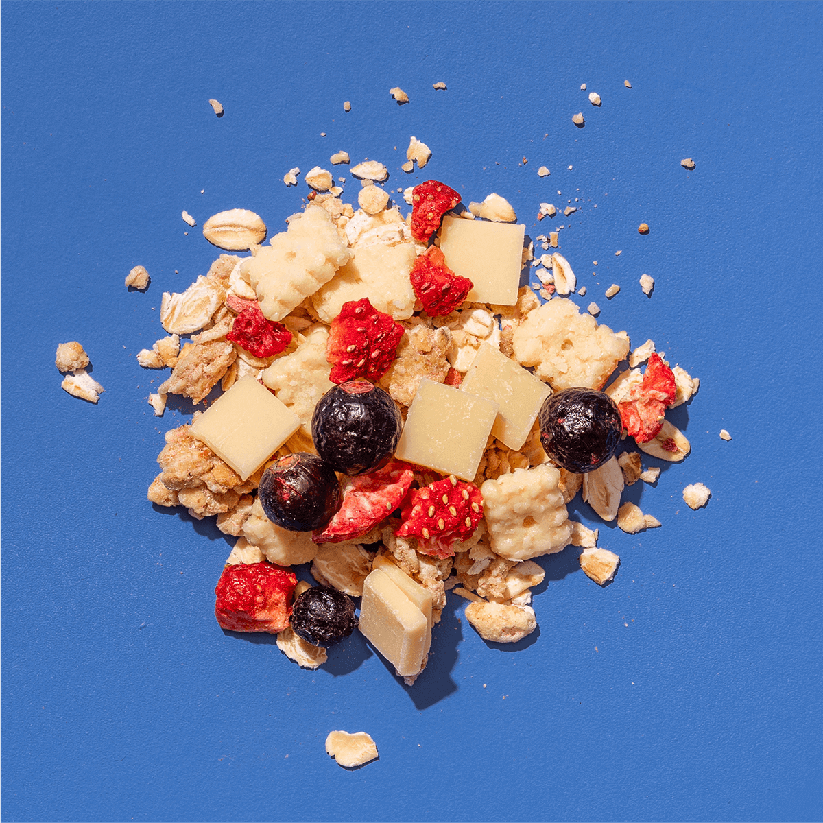 Nahaufnahme einer Müslimischung auf blauem Grund mit weißen Schokoladenstücken, Bananenchips und gefriergetrockneten Erdbeeren.