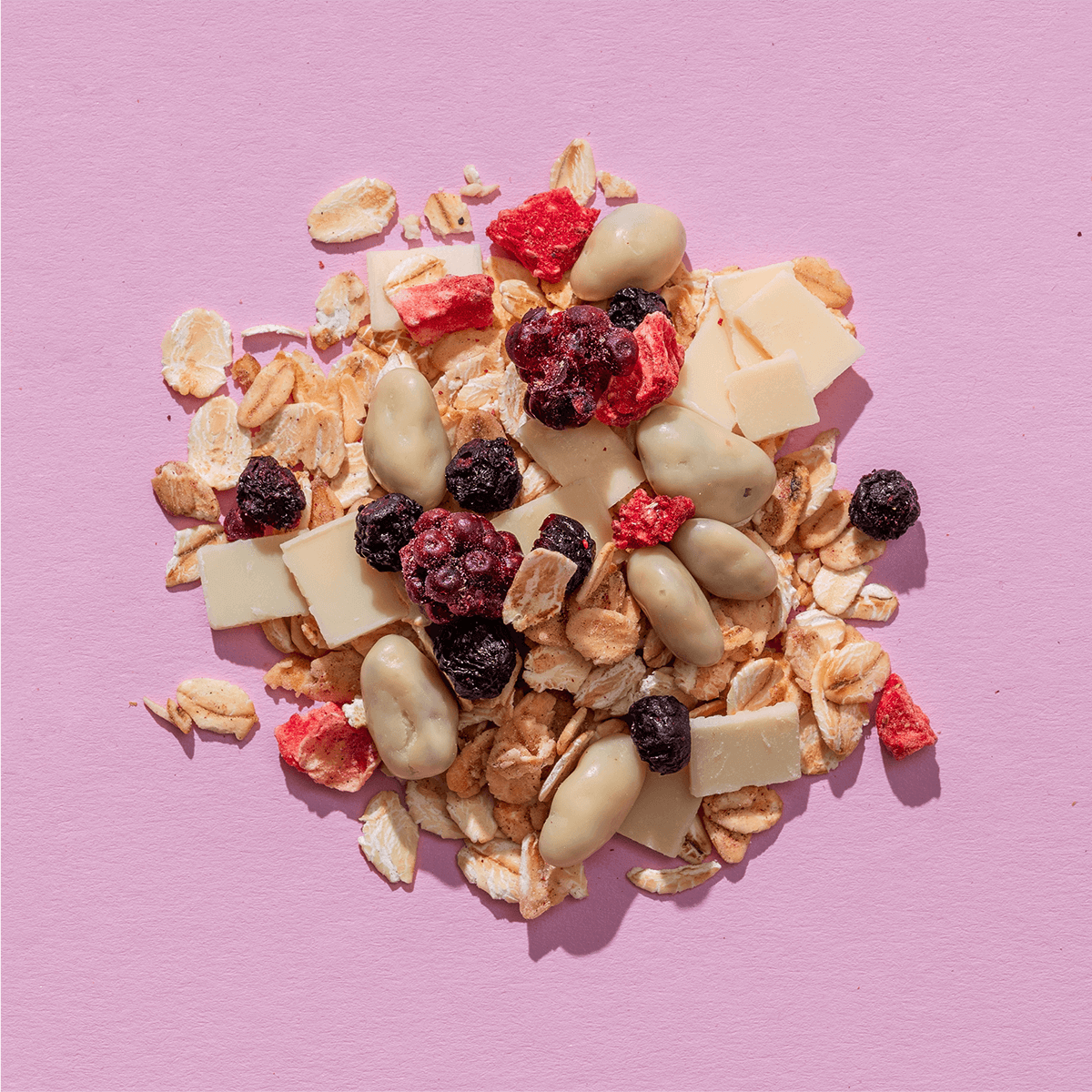 Eine Nahaufnahme einer Müsli-Mischung auf violettem Grund mit weißen Schokoladenstücken, gefriergetrockneten Beeren und Haferflocken.
