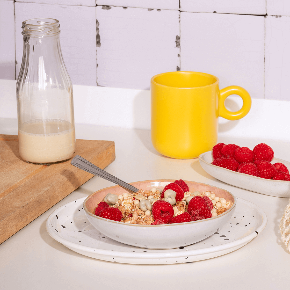 Frühstücksszene in einer hellen Küche: Eine Schale Müsli mit Himbeeren, eine gelbe Tasse und eine Glasflasche mit Milch auf einem Holzbrett.