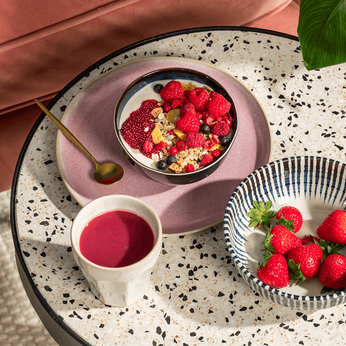 Frühstücks-Szenerie auf einer Terrazzo-Platte: Eine Schüssel Joghurt mit Beeren, eine Schale frische Erdbeeren und ein Becher mit rotem Saft.