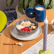 Frühstücksszene auf einem Holztisch: Eine gestreifte Schüssel mit Müsli und Früchten neben einer blauen Kaffeetasse und einer hohen lila Kerze.