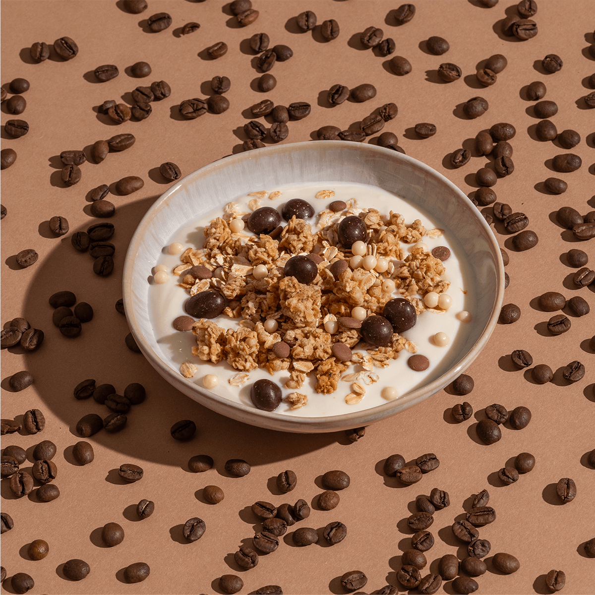 Eine Schüssel mit Müsli und Schokoladenstücken, umgeben von Kaffeebohnen auf einem braunen Untergrund.
