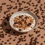 Eine Schüssel mit Müsli und Schokoladenstücken, umgeben von Kaffeebohnen auf einem braunen Untergrund.