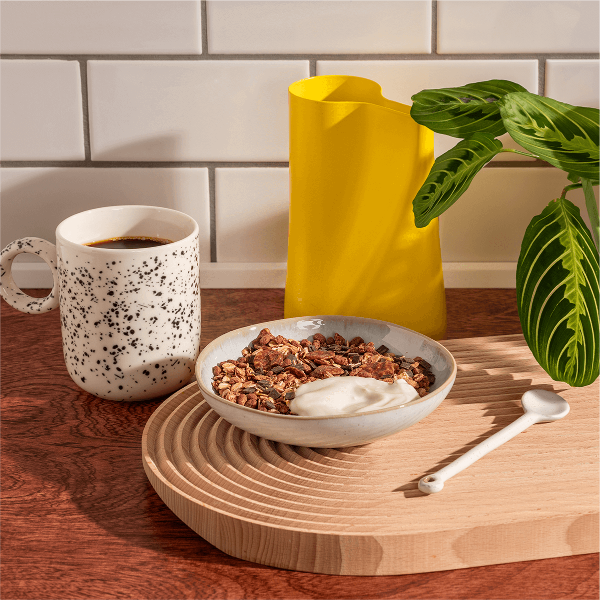 Lifestyle-Frühstücksszene mit einer Schüssel Müsli, einer Tasse Kaffee und einer gelben Kanne auf einem runden Holzbrett vor weißem Kachelhintergrund.