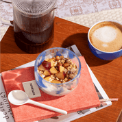 Gemütliche Frühstücksszene im Freien mit einer Müsli-Schüssel, einer French Press und einer Tasse Kaffee auf einem Holztisch.