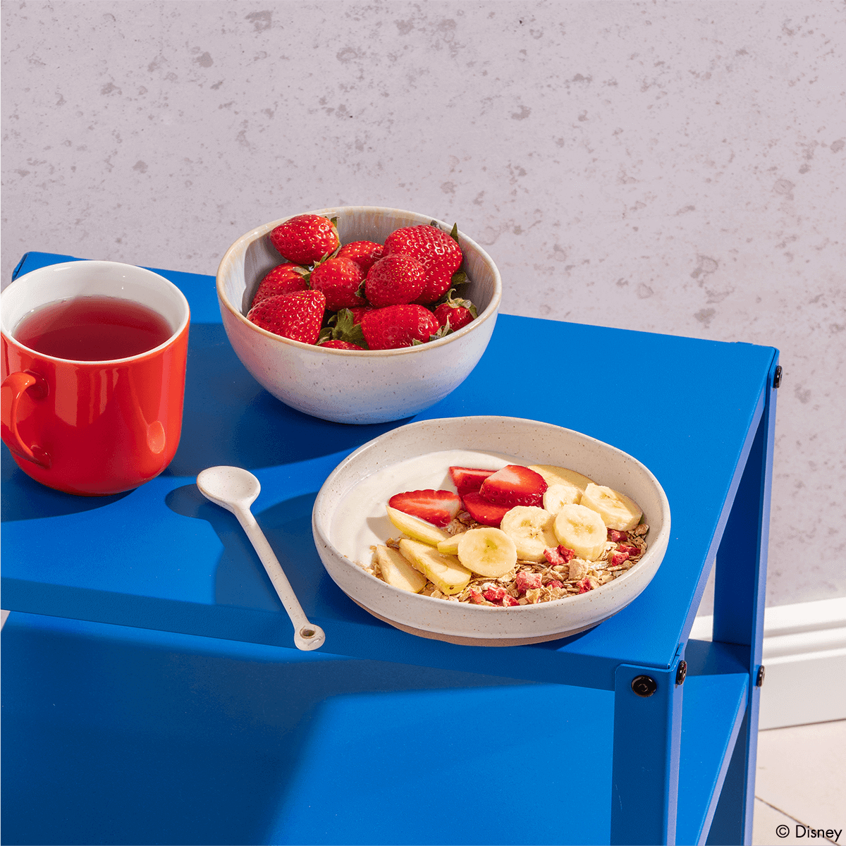 Frühstücks-Szene auf einem blauen Metalltisch: Eine Schüssel mit Müsli und Bananen, eine Schale voller frischer Erdbeeren und eine rote Tasse Tee.