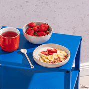 Frühstücks-Szene auf einem blauen Metalltisch: Eine Schüssel mit Müsli und Bananen, eine Schale voller frischer Erdbeeren und eine rote Tasse Tee.