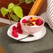 mymuesli Bio Raspberry Dark Choc müsli wird in einer Schale zusammen mit frischen Himbeeren, Pfirsichen und Yoghurt serviert. 