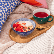 Gemütliches Frühstücks-Set auf einem Holztablett im Bett: Eine Schüssel Müsli mit Erdbeeren und eine Tasse roter Tee neben Kissen.