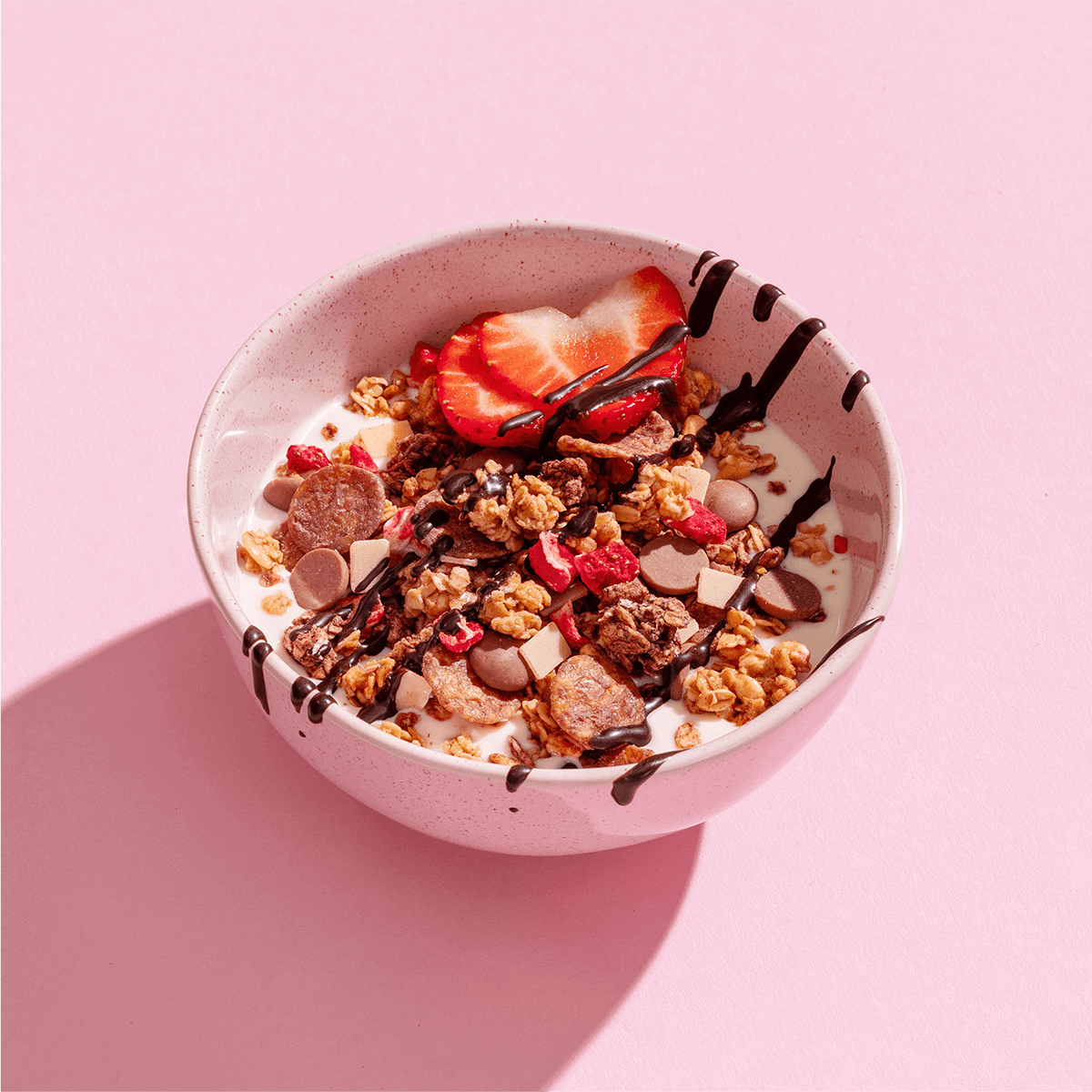 Eine Schüssel mit Müsli und Joghurt, garniert mit Erdbeerscheiben und feinen Schokoladenstreifen auf einem hellrosa Untergrund.