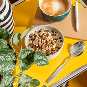 Lifestyle-Szene eines Frühstücks mit einer Schüssel Müsli, einer Tasse Kaffee und einem Notizbuch auf einem gelben Tisch.