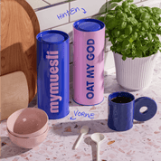 Zwei Vorratsdosen blau & rosa stehend neben einer Tasse und Schalen