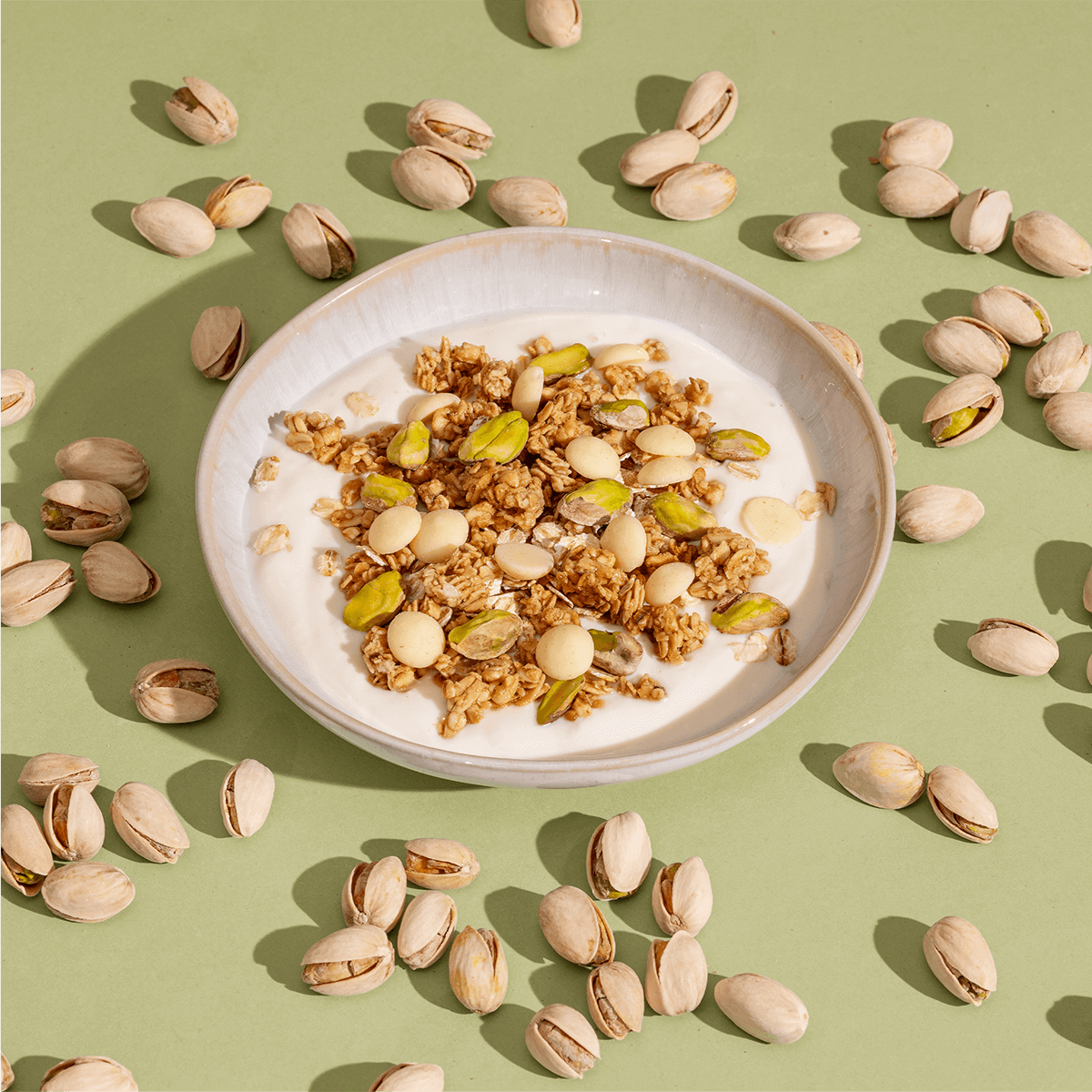 Eine Schüssel mit Joghurt und Pistazien-Granola, umgeben von Pistazien in der Schale auf einem lindgrünen Hintergrund.