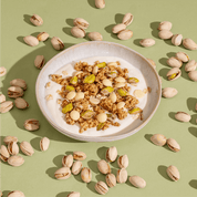 Eine Schüssel mit Joghurt und Pistazien-Granola, umgeben von Pistazien in der Schale auf einem lindgrünen Hintergrund.