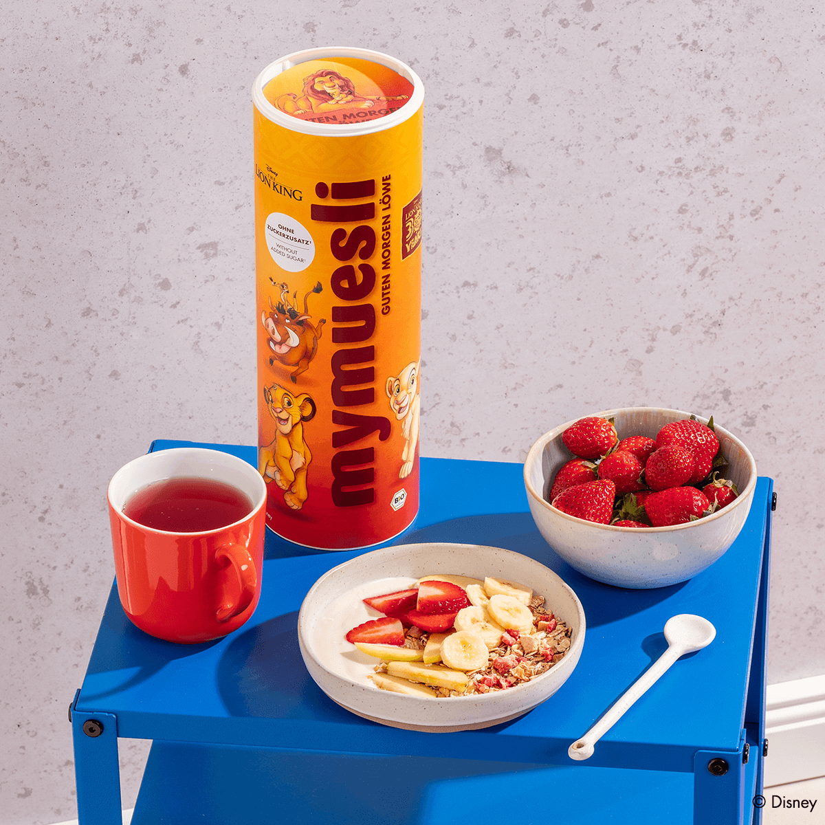 Eine gelbe mymuesli -Dose (König der Löwen Edition) steht auf einem blauen Beistelltisch neben einer Müsli-Bowl, einer Schale Erdbeeren und einer roten Tasse.